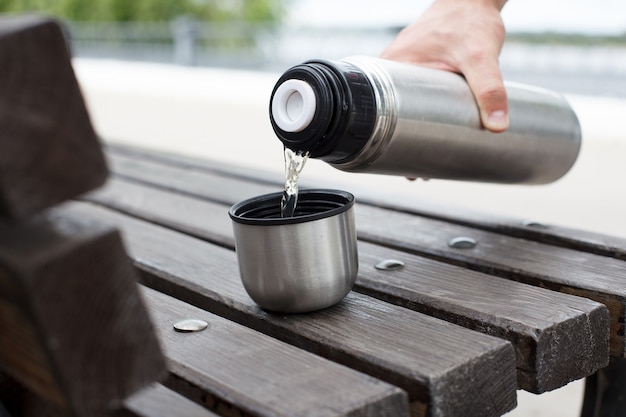 La mano de un hombre vierte té de un termo en una taza en un banco.