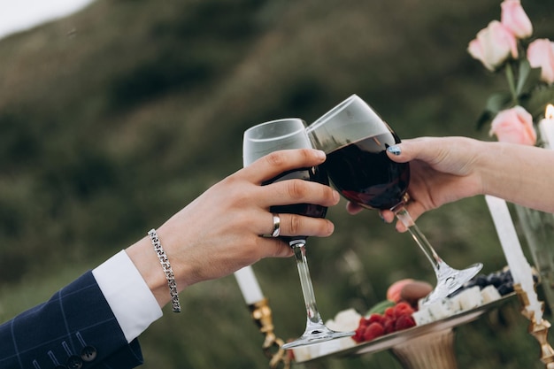 Mano de hombres y mujeres sostienen copas de vino tinto Fecha romántica a la luz de las velas al aire libre Copas de tintineo primer plano