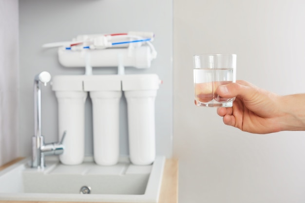 La mano de los hombres sostiene un vaso de agua clara. Toque y filtro de ósmosis inversa en el fondo.