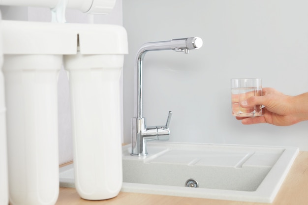 La mano de los hombres sostiene un vaso de agua clara. Toque y filtro de ósmosis inversa en el fondo.