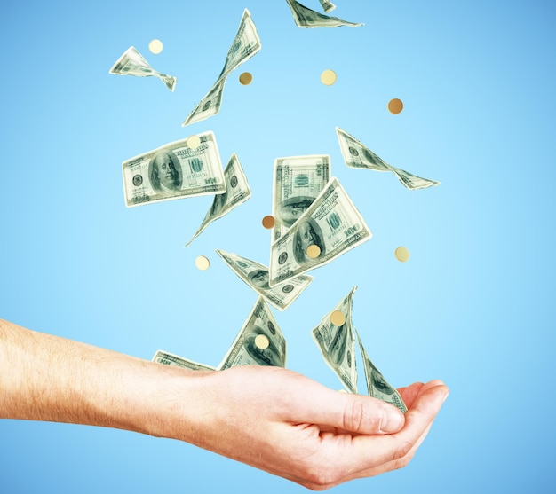 Mano humana con dinero y monedas cayendo sobre fondo azul.