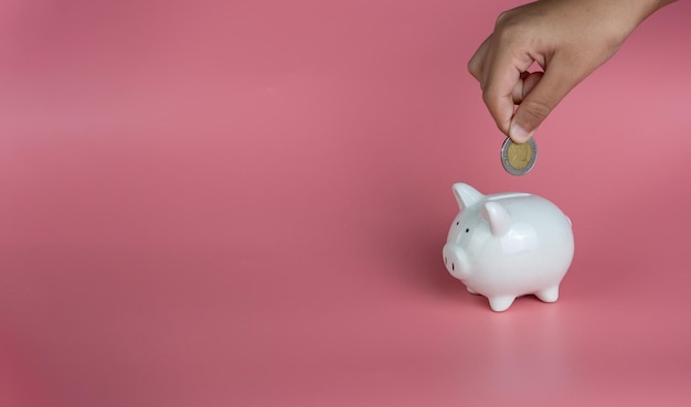 Una mano humana está dejando caer una moneda en una alcancía blanca sobre un fondo rosa Ahorros.