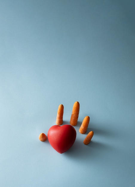 Foto una mano humana hecha de zanahorias frescas sale del fondo azul y alcanza el corazón rojo. concepto de salud y autocuidado. concepto mínimo creativo.