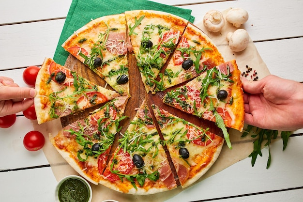 La mano humana toma pizza recién horneada con pesto de salsa rukkola de jamón y aceitunas servidas sobre fondo de madera con salsas de tomate y hierbas Concepto de entrega de alimentos