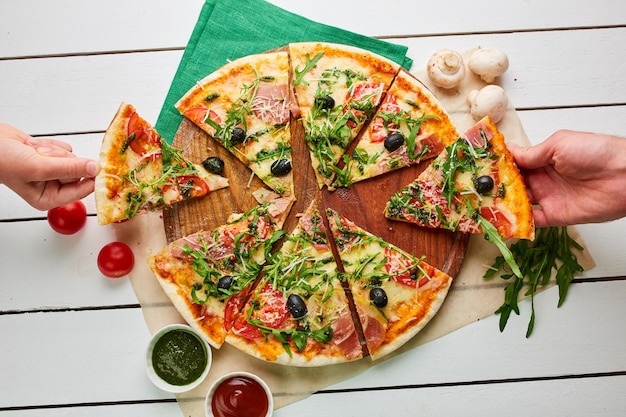 La mano humana toma pizza recién horneada con pesto de salsa rukkola de jamón y aceitunas servidas sobre fondo de madera con salsas de tomate y hierbas Concepto de entrega de alimentos