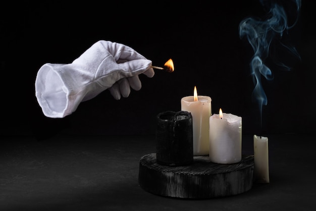 Mano invisible en un guante blanco encendiendo velas con un fósforo, humo de velas sobre un fondo negro, concepto aterrador de halloween.