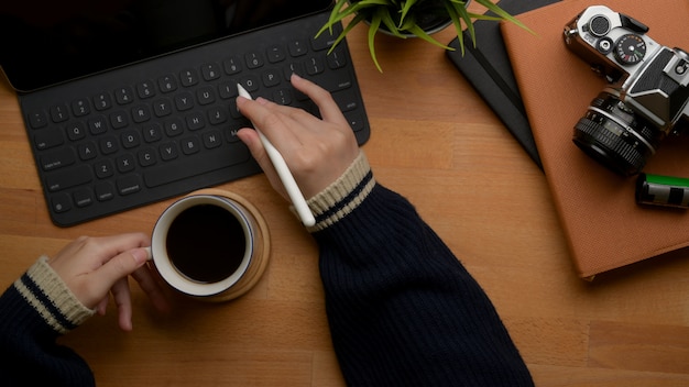 Mano izquierda femenina sosteniendo la taza de café y la mano derecha escribiendo en el teclado de la tableta sobre la mesa de trabajo