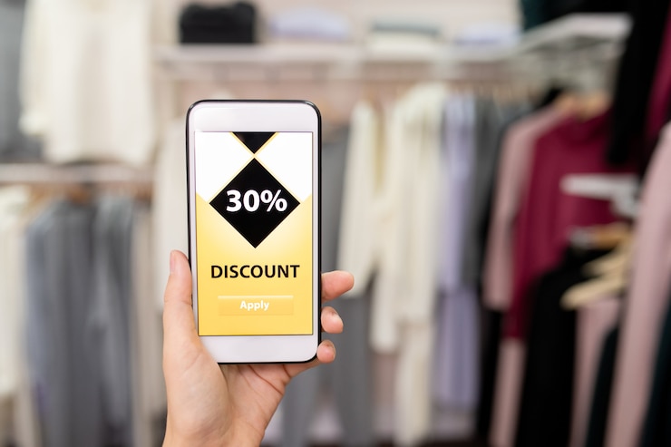 Foto mano de joven adicta a las compras femenina contemporánea sosteniendo smartphone con cupón de descuento en su pantalla mientras va a comprar ropa en boutique