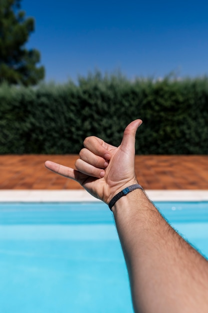 Mano del joven caucásico mostrando los dedos sobre fondo azul borroso piscina gesticulando gesto de saludo shaka hawaiano, símbolo de teléfono y comunicación