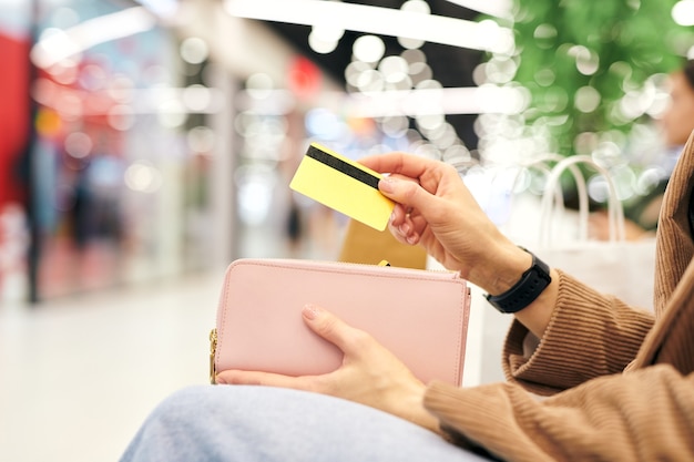 Mano de joven clienta contemporánea poniendo una tarjeta de plástico en la billetera mientras descansa en el entorno del centro comercial después de ir de compras