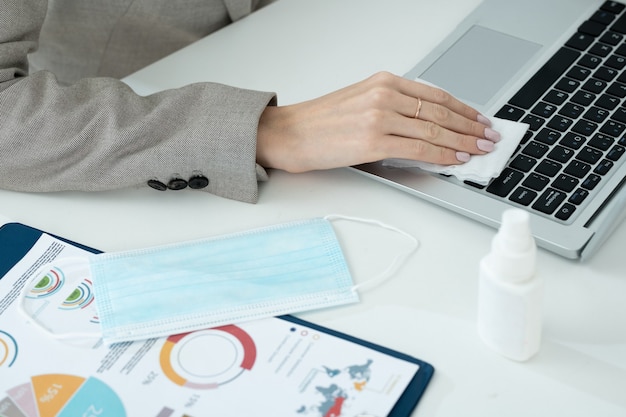 Mano de joven empresaria en ropa formal gris limpiando el teclado de la computadora portátil con un paño empapado con desinfectante mientras está sentado en el lugar de trabajo en la oficina