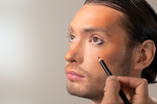 Foto mano del joven maquillador con delineador de ojos kohl negro que se va a aplicar en los ojos del modelo masculino