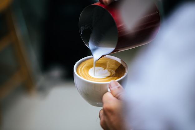 La mano de la leche de colada del barista hace arte del latte del café con la taza blanca