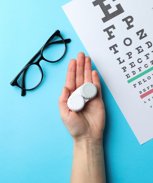 Mano con lentes de contacto, anteojos y tabla de examen ocular en superficie azul, vista superior