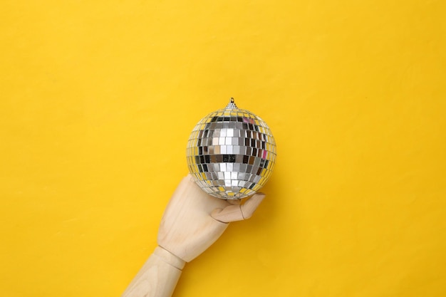 Mano de madera que sostiene la bola de discoteca sobre fondo amarillo Vista superior Concepto de fiesta minimalista endecha plana