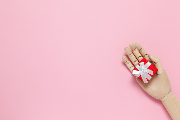 La mano de madera sostiene una pequeña caja de regalo roja con lazo blanco