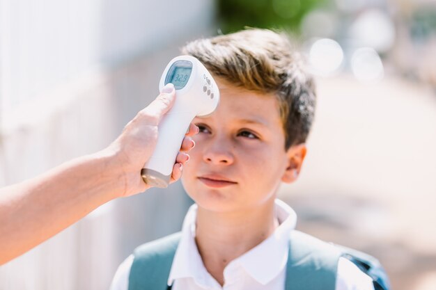 Mano del maestro tomando la temperatura de un niño al ingresar a la escuela con un termómetro electrónico, con la cabeza del estudiante desenfocada. Covid-19 y concepto de pandemia de virus