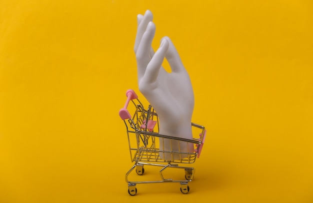 Mano de maniquí blanco en carrito de compras sobre fondo amarillo.