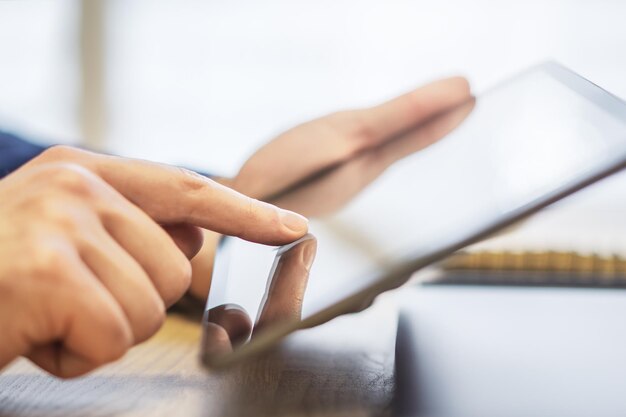 Foto mano masculina interactuando con un dispositivo de tableta digital de vanguardia que descansa sobre un escritorio contra el ajetreo desenfocado de una oficina iluminada por el sol