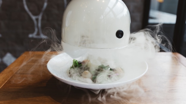 La mano masculina se levanta para levantar la campana de vidrio de un plato con comida caliente y humo en movimiento en el restaurante.