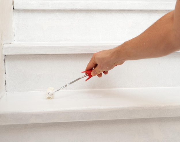 Mano masculina pinta una escalera de madera blanca con pintura y un rodillo