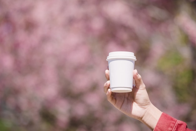 Mano masculina que aparece de una taza blanca de café caliente delante de flor de cerezo rosa