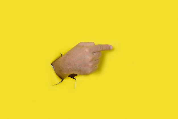 Foto mano masculina saliendo de un agujero en un fondo de papel roto amarillo apuntando a la derecha