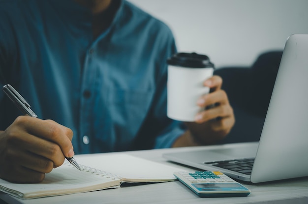 Mano masculina sosteniendo un bolígrafo y escribiendo en un cuaderno y una taza de café en el escritorio con una computadora portátil y una calculadora