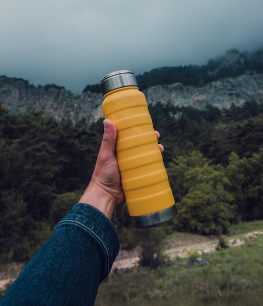 Mano masculina sosteniendo una botella térmica amarilla de acero en el fondo de la naturaleza