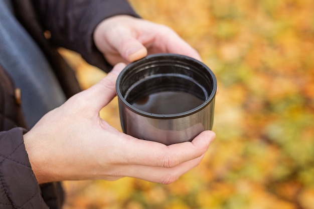 Mano masculina sosteniendo una taza termo con té negro caliente en el bosque de otoño - Imagen