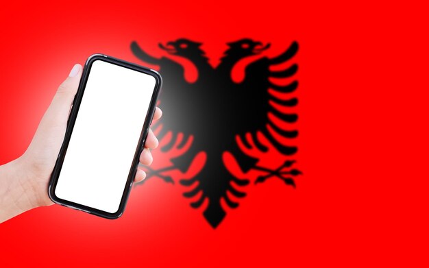 Mano masculina sosteniendo el teléfono inteligente con pantalla en blanco en el fondo de la bandera borrosa de Albania Vista de cierre