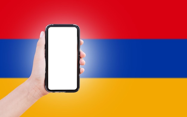 Mano masculina sosteniendo el teléfono inteligente con pantalla en blanco en el fondo de la bandera borrosa de Armenia Vista de cierre