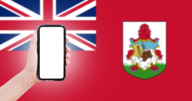 Mano masculina sosteniendo el teléfono inteligente con pantalla en blanco en el fondo de la bandera borrosa de las Bermudas Vista de cierre