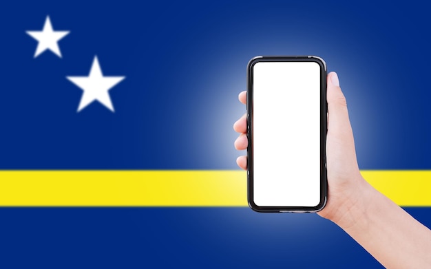 Mano masculina sosteniendo el teléfono inteligente con pantalla en blanco en el fondo de la bandera borrosa de Curacao Vista de cierre