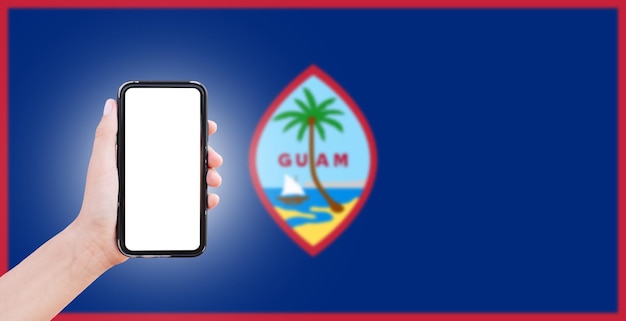 Mano masculina sosteniendo el teléfono inteligente con pantalla en blanco en el fondo de la bandera borrosa de Guam Vista de cierre