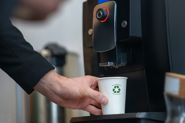 Mano masculina sosteniendo un vaso de papel blanco con letrero verde cerca del soporte en la máquina de bebidas