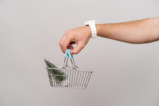 Mano masculina sostiene una cesta de la compra con un pequeño árbol de Navidad en ella