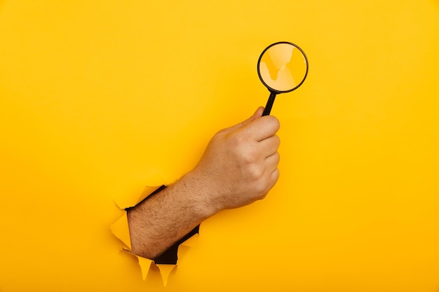 La mano masculina sostiene una lupa negra en un agujero rasgado de fondo amarillo Concepto de búsqueda de información