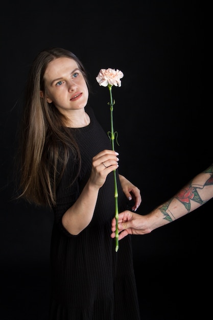 Mano masculina con tatuajes da flor de clavel a hermosa chica morena con cabello largo, concepto de felicitación sobre fondo negro