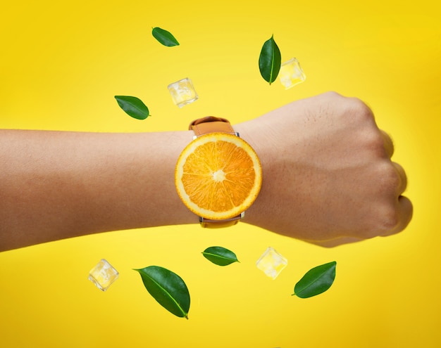 Mano masculina vistiendo naranja de frutas Reloj Hojas verdes y cubos de hielo volando