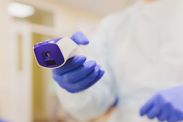 La mano del médico en un guante protector sostiene un termómetro infrarrojo y mide la temperatura del paciente. Medición remota de fiebre alta para un paciente con coronovirus durante una pandemia.