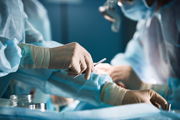 Mano del médico con guantes de látex con pinzas de acero inoxidable para ayudar a las personas con paciencia, sobre un fondo gris blanco.