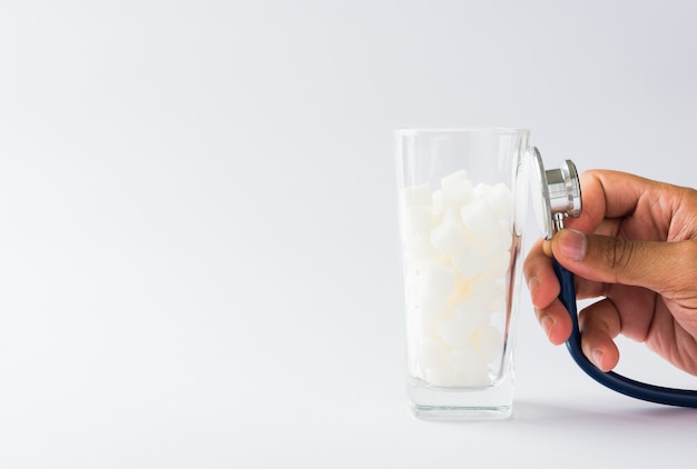 La mano del médico mantenga el control del estetoscopio en el vaso lleno de ingrediente alimentario dulce de terrón de azúcar blanco