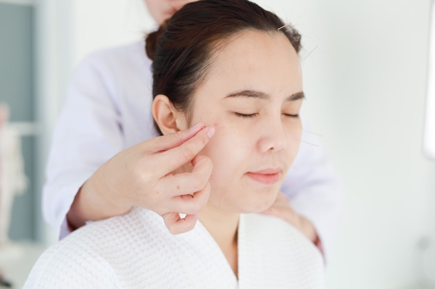 Mano del médico que realiza la terapia de acupuntura. Mujer asiática sometida a tratamiento de acupuntura con una línea de agujas finas insertadas en la piel de su cara en un hospital clínico