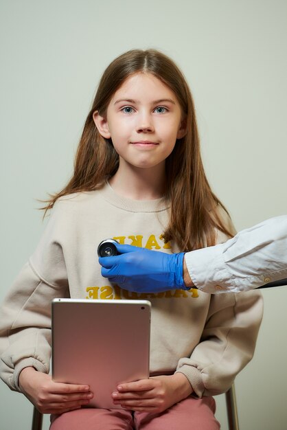 La mano de un médico sostiene un estetoscopio escuchando los latidos del corazón de una paciente joven. Una muchacha feliz con una sonrisa que sostiene una tableta en la cita del doctor.