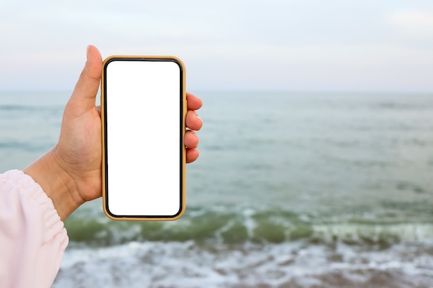 Mano mostrando un teléfono inteligente en blanco en la playa con el mar de fondo. Maqueta de pantalla blanca