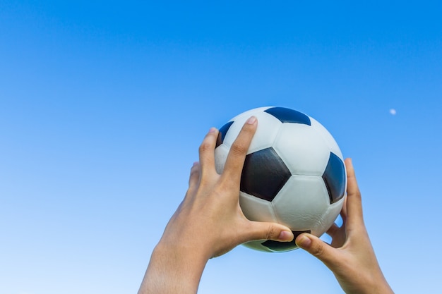 La mano de la muchacha sostiene un balón de fútbol en el cielo en fondo del cielo azul.