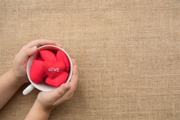 La mano del muchacho sostiene los corazones rojos en la taza de café en fondo de la textura del saco