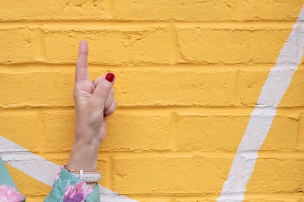 Una mano de mujer apuntando hacia arriba con el dedo índice contra una pared de ladrillo amarillo