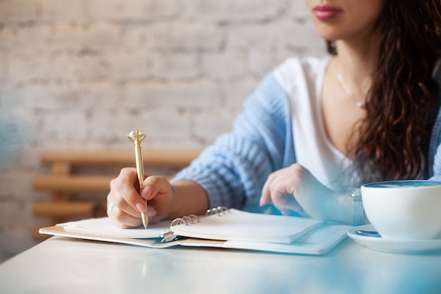 Mano de mujer bien arreglada sosteniendo bolígrafo dorado y escribiendo notas con bolígrafo dorado en el cuaderno mientras bebe café con leche azul junto a la ventana. Periodista autónomo que trabaja desde casa. Planificación del concepto futuro. Copia espacio